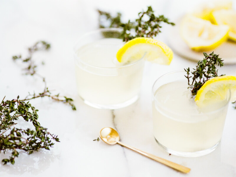 Two white cocktails in small tumblers sit on a marble worktop. The glasses are decorated with fresh thyme.